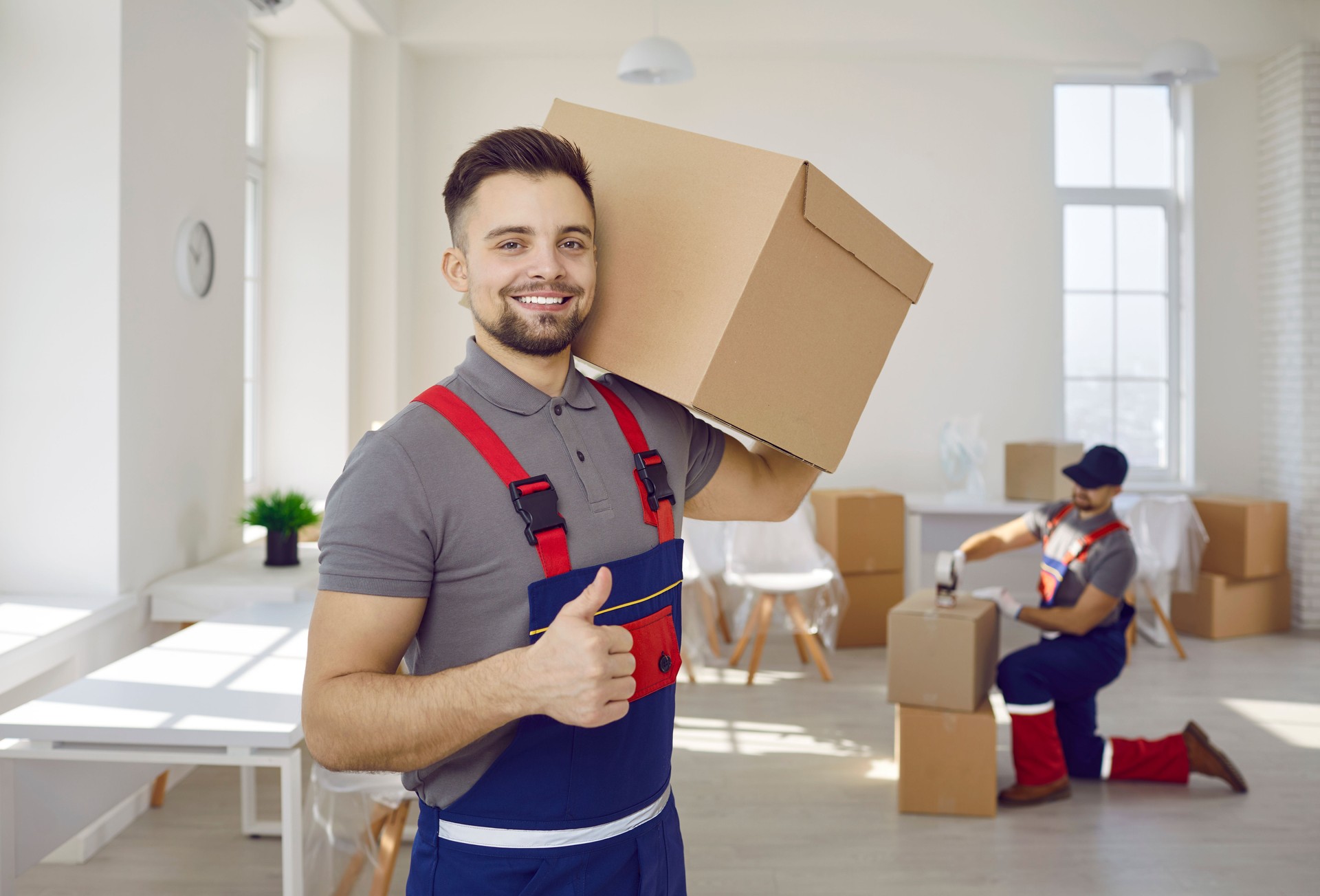 Trabajador masculino sonriente de una empresa de mudanzas y entregas sosteniendo una caja de cartón mostrando los pulgares hacia arriba.